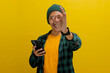 A young Asian man, dressed in a beanie hat and casual clothes, makes an OK sign gesture while holding his smartphone, conveying agreement, approval, and satisfaction, indicating a positive review
