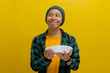 Hungry young Asian man looks towards an empty copy space while holding a plate in his hand, excitedly contemplating what to eat, as he stands against a yellow background