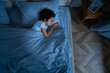 © Prostock-studio - A young African American boy is laying comfortably in bed with a soft blue comforter covering him. He appears relaxed and at ease, possibly getting ready to sleep or just waking up, view above