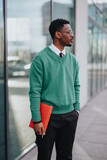 Fototapeta  - Professional male entrepreneur with eyeglasses and green pullover stands outside a modern building, clutching a bright red folder.