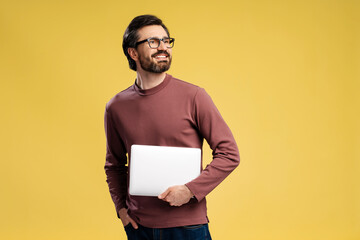 Happy young man in eyeglasses with laptop posing in studio, isolated on yellow