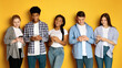 © Prostock-studio - A diverse group of five multiethnic teenagers stand side-by-side against a vibrant yellow backdrop, each engrossed in their own smartphone