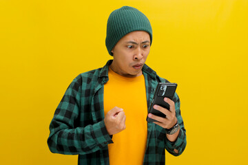 Wall Mural - An angry Asian man, dressed in a beanie hat and casual shirt, visibly reacts to bad news while looking at his mobile phone, while standing against yellow background