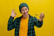 Excited young Asian man, dressed in a beanie hat and casual shirt, samples food with a spoon, giving an OK hand gesture, smiling at the camera, expressing satisfaction with the taste yellow background