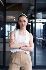 Young Caucasian female in casual office attire stands, arms crossed in modern office