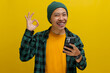 A young Asian man, dressed in a beanie hat and casual clothes, makes an OK sign gesture while holding his smartphone, conveying agreement, approval, and satisfaction, indicating a positive review