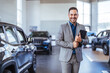 Cropped portrait of a handsome young male car salesman working on the showroom floor. Happy salesperson standing in a car showroom looking at camera.
