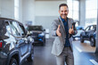 Good looking, cheerful and friendly salesman poses in a car salon or showroom. A professional car salesman in a suit holding a tablet in a car dealership showroom.