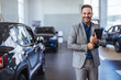 Salesperson at car dealership selling vehicles. Front view of handsome man standing in modern car center and posing. Bearded manager smiling and looking at camera.