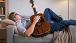 Musician relax. Home rest. Peaceful casual dreamy man laying on sofa playing acoustic guitar composing melody in light cozy living room interior.
