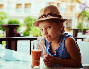 Wall Mural - Thinking kid girl looking serious in summer hat drinking and drinking tasty healthy juice in street restaurant. Closeup