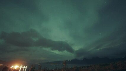 Wall Mural - Mesmerizing display if the northern lights in the night sky above the dark waters of the fjord and brightly illuminated port.