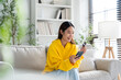 Young asian woman sitting on sofa in living room, makes online banking payments through the internet from bank card on cell phone. Shopping online on mobile with credit card