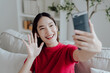Excited happy young Asian woman holding smart phone device sitting on sofa at home - Happy satisfied female looking at mobile smartphone screen and gesturing greeting friend with video call