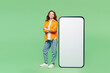 © ViDi Studio - Full body happy young woman wear orange shirt white t-shirt casual clothes stand near big huge blank screen mobile cell phone smartphone with area isolated on plain green background Lifestyle concept