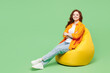 © ViDi Studio - Full body happy cheery pretty young ginger woman wear orange shirt white t-shirt casual clothes sit in bag chair rest look camera isolated on plain pastel light green background. Lifestyle concept