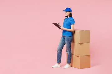 Sticker - Full body delivery employee woman she wear blue cap t-shirt uniform work dealer courier hold clipboard papers document, stack of clear cardboard box isolated on plain pink background. Service concept.