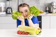 Cute little girl with lettuce leaves and a plate of vegetables with tomatoes and celery sitting at the table in the kitchen. Selected focus.
