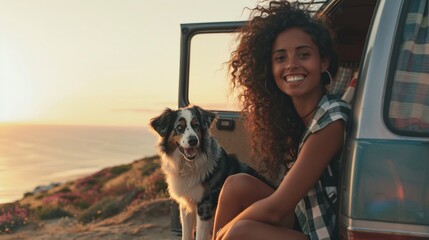 a woman with long curly hair sits on the back of a van, smiling as her black and white dog sits beside her the van has a window and the woman's bare leg