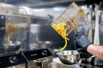 Wall Mural - Hands in black gloves pouring fried potatoes from a metal fryer basket into a stainless steel bowl