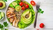 A nutritious salad with grilled chicken, quinoa, avocado slices, and cherry tomatoes on white wooden background