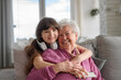 Cute girl hugging gradmother from behind, looking at camera, smiling. Portrait of an elderly woman spending time with granddaughter.