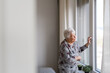 Senior woman looking out of window, spending time alone in her apartment. Concept of loneliness and dependence of retired people.