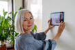 Mature woman adjusting smart thermostat, touch screen of smart home device. Older woman using smart technology at home.