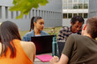 multiracial group of university students sitting on a bench in the campus gardens, with laptops and academic material doing a class assignment together.