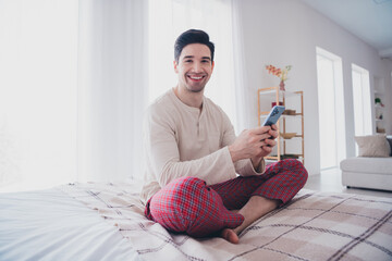 Poster - Photo of cheerful domestic man sitting on comfy bed weekend day white light room interior indoors