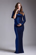 Portrait of young pretty pregnant woman on gray studio background. Female in blue sequin dress with hands near pregnant belly