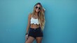 © VISUAL BACKGROUND - A woman in black shorts and white crop top posing against a blue wall.
