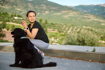 Wall Mural - Smiling happy woman sitting with her dog in the mountains, enjoying walk and spending time with her pedigree black cocker spaniel pet, dreamily looking aside into the distance. People and playing pets