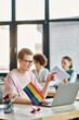 © Bliss - Short haired woman engrossed in work, with a laptop in front of her, with her diverse colleagues on backdrop, pride flag.