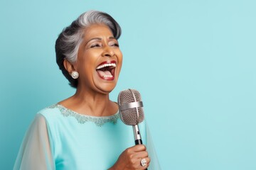 Wall Mural - Portrait of a grinning indian woman in her 50s dancing and singing song in microphone while standing against pastel blue background