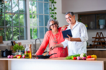 Indian asian senior old age cheerful couple using tablet computer or laptop in the kitchen while cooking