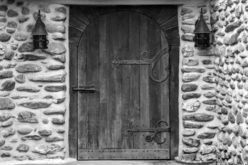 The stone wall of an old house with lamps and a wooden door with forged elements. Beautiful fabulous house, fairytale. Copy space. Black and white photo. Selective focus