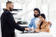 © Prostock-studio - A young Indian couple is in the process of purchasing a new car, as they stand signing documents presented by a friendly salesperson inside a bright car dealership showroom