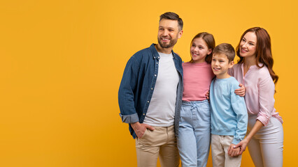 A happy family, with two parents and two children, embrace each other against a vibrant yellow backdrop. They appear cheerful and content, looking at copy space