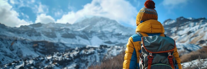 Wall Mural - A boy in a colorful jacket, a cap with a bubo and a backpack on his shoulders against the backdrop of a snowy mountain and a winter blue sky, Back view realistic nature and landscape