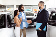 © Prostock-studio - A young Indian couple is finalizing a deal with a car salesman inside a vehicle showroom. They seem pleased and are shaking hands with the agent, who is dressed in a business suit