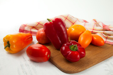 Cutting board with several red and yellow tomatoes and bell pepper on white wooden background with red kitchen towel..