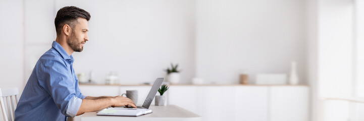 Poster - Handsome millennial businessman sitting at workplace in white office, working on laptop, typing on computer keyboard, answering emails, corresponding with clients, side view with copy space