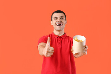 Wall Mural - Young man holding french fries and showing thumb-up gesture on orange background