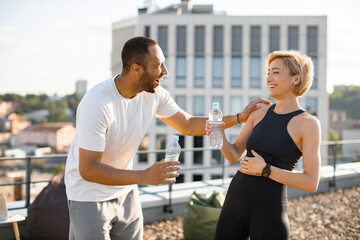 Wall Mural - Athletic man in white t-shirt and woman in black top drinking water after jogging. Young couple having fun laughing after morning workout on the roof of the house.