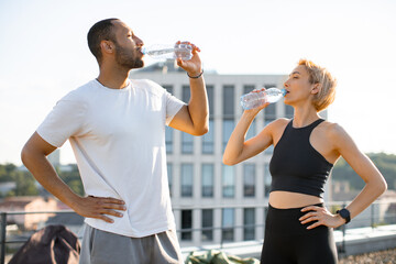 Wall Mural - Young athletic family engaged in healthy lifestyle during morning warm-up and workout on roof of modern building. Athletic sporty man and fit woman drinking water after jogging outdoors.