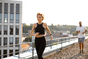 Young sporty woman in black top running in morning with male partner in white t-shirt against urban morning background. Concept of healthy lifestyle and positive start of day.