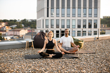 Wall Mural - Young couple in sportswear sitting on mats in lotus position on roof of modern house. Athletic man and flexible woman meditating in morning on urban background.