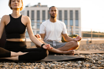 Wall Mural - Close up view of hands on knees of young couple in sportswear sitting on mats in lotus position on roof of modern house. Athletic man and flexible woman meditating in morning on urban background.