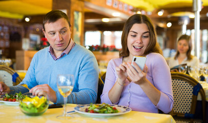 Wall Mural - Modern couple having lunch in cozy restaurant, sitting at table and using mobile phones. Phubbing concept..